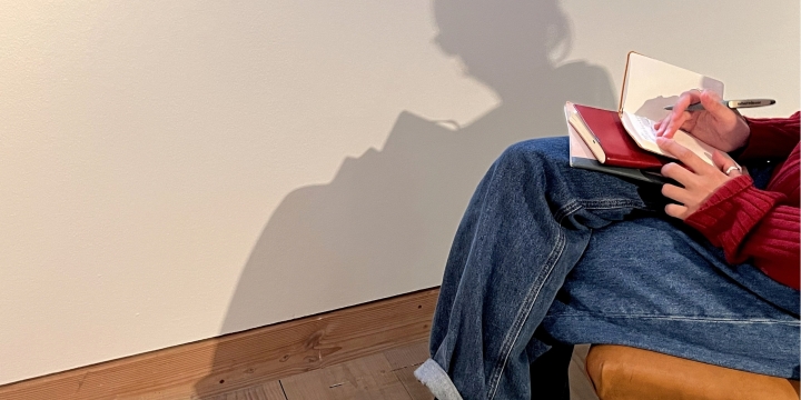 sitzende Person, mit Schreibblock und Stift auf den Knien auf einem Stuhl auf Holzboden in einem Raum mit weißer Wand