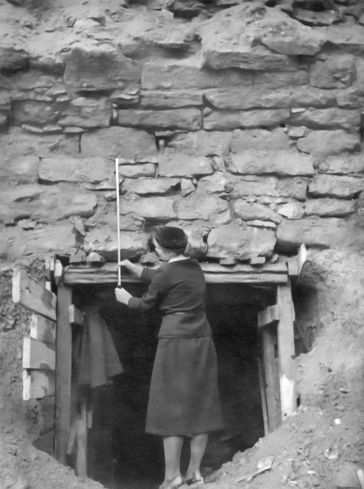 Frau mit Maßstab vor vor einer ausgegrabenen Mauer mit Eingang