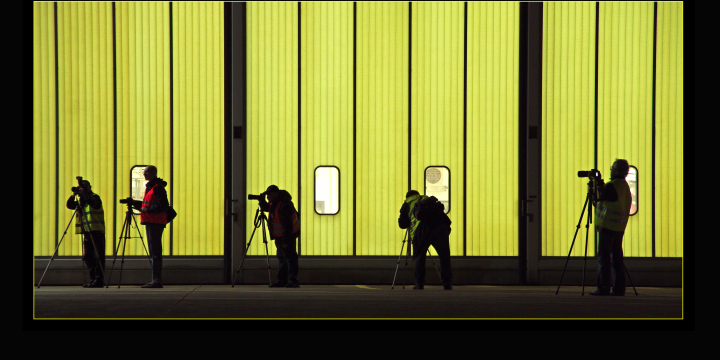 Silhouetten fotografierende Personen vor einer hinterleuchteten gelben Fensterfront